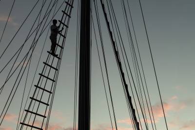 A silhouetted figure climbs a rope ladder against a mast, with a soft pink and gray sky in the background.