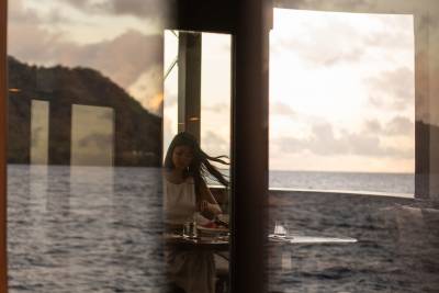 A woman dines at a table reflected in a window overlooking the ocean at sunset.