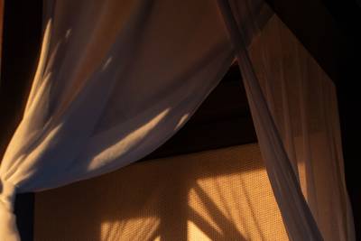 A sheer white curtain in the foreground is illuminated by warm sunlight, casting shadows onto a textured woven background.