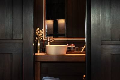 A modern sink with a brass faucet sits on a wooden vanity in the foreground, while a mirror reflects a dimly lit room in the background.