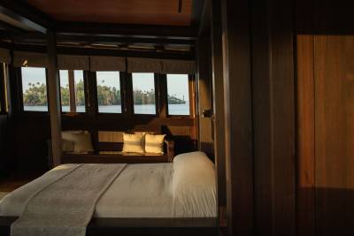 A neatly made bed with white linens and a light gray throw occupies the foreground, while a serene body of water with lush green trees is visible through the windows in the background.