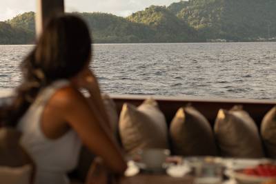 A woman in a white dress looks out at a calm sea with lush green hills in the background, from a comfortable seating area with plush pillows.
