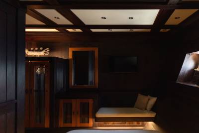 A dimly lit room features a wooden wardrobe and mirror in the foreground, with a built-in bench and a television on the dark wood paneled wall in the background.