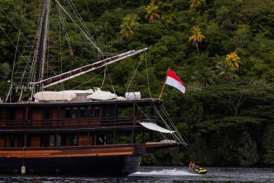 A large wooden boat with an Indonesian flag sails past a lush, green, tropical hillside where a jet ski speeds across the water.