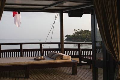A daybed with pillows and a tray sits on a wooden deck in the foreground, while a calm ocean and a lush island are visible in the background under a pale sky.