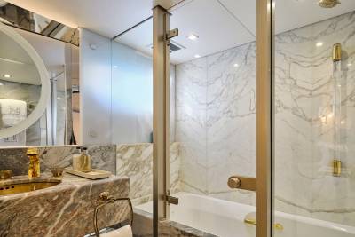A luxurious bathroom features a marble vanity with a gold faucet in the foreground and a marble-tiled shower and bathtub in the background.