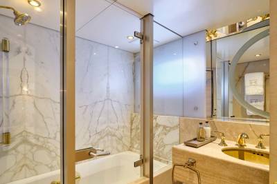 A luxurious bathroom features a marble bathtub and shower in the foreground, with a vanity and mirror reflecting a towel in the background.