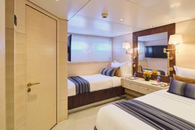 Two neatly made beds with striped pillows sit in a well-lit cabin, with a large mirror reflecting a television and lamps on the wall behind them.