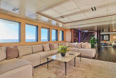 A spacious, modern yacht lounge with a large sectional sofa and coffee table in the foreground, and a staircase and dining area visible in the background.
