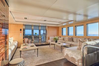 A luxurious yacht lounge with plush seating and coffee tables in the foreground opens to a sunlit deck with ocean views in the background.