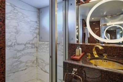 A luxurious bathroom features a marble shower stall in the foreground and a gold-accented sink with a backlit mirror in the background.
