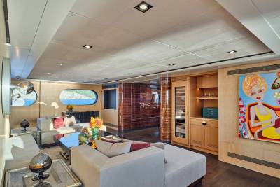 A modern yacht interior features a white sectional sofa and colorful artwork in the foreground, with oval windows revealing a coastal landscape in the background.