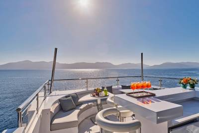 A luxurious yacht deck with a curved sofa and bar in the foreground overlooks a serene ocean and distant mountains under a bright sun.