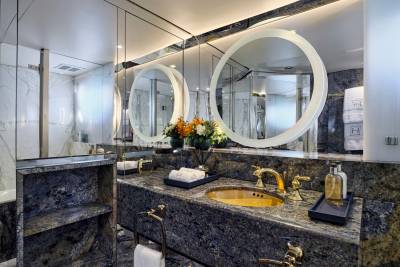 A luxurious bathroom features a granite countertop with a gold faucet in the foreground, while the background showcases large, illuminated mirrors reflecting the elegant marble walls and shower.