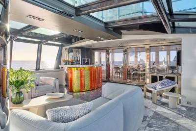 A luxurious yacht interior with plush seating in the foreground and a bar and dining area visible through glass doors in the background, all under a glass ceiling with ocean views.