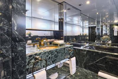 A luxurious bathroom with a green marble vanity and sink in the foreground, and a large mirrored wall reflecting the bathtub and cabinetry in the background.