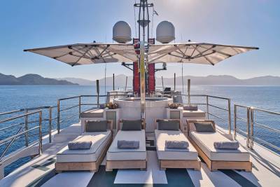 Four sun loungers with grey cushions are arranged on the deck of a yacht, with a clear blue sky and distant, hazy mountains in the background.