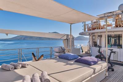 Rolled towels and plush cushions adorn the sun loungers on the deck of a yacht, with a calm blue sea and distant islands forming the backdrop.