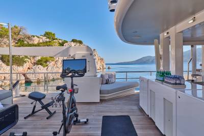 A Peloton bike and weights are set up on a yacht's deck with a scenic coastal view in the background.