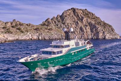 A green yacht cruises through blue water in the foreground, with a rocky, sunlit island in the background under a clear sky.