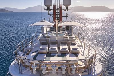 Sun loungers and a hot tub are arranged on the deck of a yacht in the foreground, with calm blue water and distant, hazy mountains in the background.