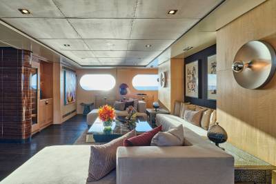 A luxurious yacht interior features a plush sectional sofa and coffee table in the foreground, with oval windows revealing a serene ocean view in the background.