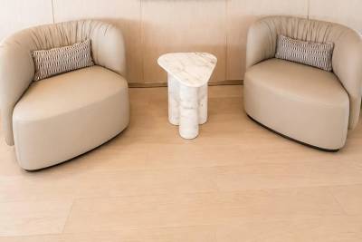 Two plush cream armchairs with decorative pillows flank a marble-topped side table on a light wood floor, with a textured wall in the background.