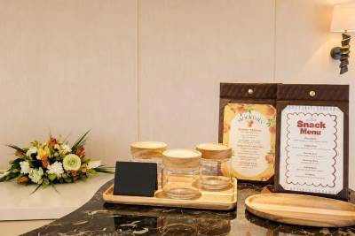 A wooden tray with jars and a small chalkboard sits on a marble counter in front of two menus, with a floral arrangement to the left and a wall with a lamp to the right.