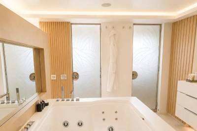 A luxurious bathroom features a large white jacuzzi tub in the foreground, with frosted glass shower doors and wooden accent walls in the background.