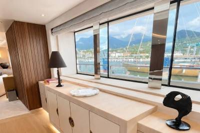 A modern interior with a wooden accent wall and a sleek cabinet in the foreground overlooks a scenic harbor with boats and mountains in the background.