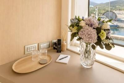 A wooden tray with glasses sits on a desk in front of a window overlooking a harbor, with a vase of flowers and a notepad on the desk.