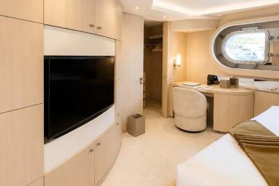 A modern bedroom with a large television and built-in cabinets in the foreground, and a desk with a porthole window in the background.