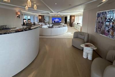 A curved white bar dominates the foreground, leading to a spacious, modern living area with a circular sofa and a large screen TV in the background.