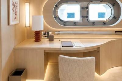 A modern desk with a lamp and laptop sits in the foreground, while a large oval window with two portholes looks out onto a bright, blue background.