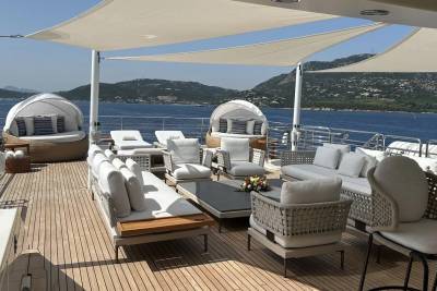 A luxurious yacht deck with comfortable seating and shaded lounging areas is set against a backdrop of a calm blue sea and distant, verdant hills.