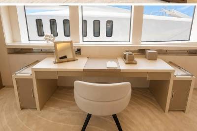 A white vanity desk with a mirror and chair sits in the foreground, while a large white yacht with multiple windows is visible through the windows in the background.