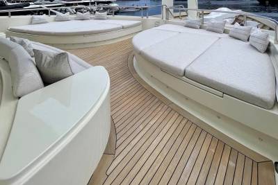 Plush, cream-colored sunbeds with pillows rest on a polished wooden deck in the foreground, while a harbor with docked boats is visible in the blurred background.