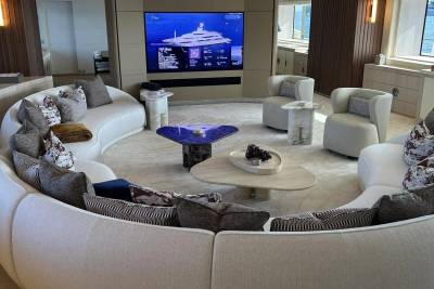 A large, curved white sofa with patterned pillows surrounds a blue, triangular coffee table in the foreground, while a large screen displaying a yacht hangs on the wall in the background.