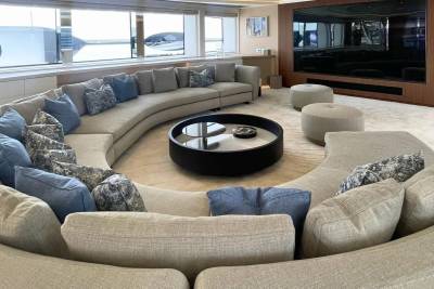 A circular sectional sofa with blue and patterned pillows surrounds a dark round coffee table in the foreground, while a large television and minimalist decor are visible in the background.