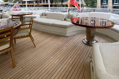 A polished wooden deck with chairs and tables in the foreground leads to a luxurious yacht with seating and flags in the background.