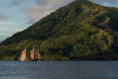 A wooden sailing ship with reddish-brown sails glides across dark blue water in the foreground, with a lush green, tree-covered mountain rising steeply in the background.