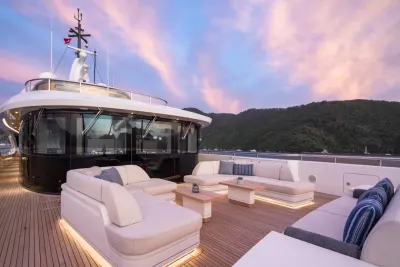Luxurious white sofas and tables adorn the wooden deck of a yacht in the foreground, while a lush, green, mountainous coastline is visible in the background under a pastel-colored sky.