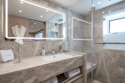 A modern bathroom with a marble vanity and sink in the foreground, and a glass-enclosed shower with a window in the background.