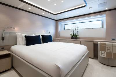A luxurious yacht bedroom features a large bed with navy pillows in the foreground and a long counter with a window overlooking the sea in the background.