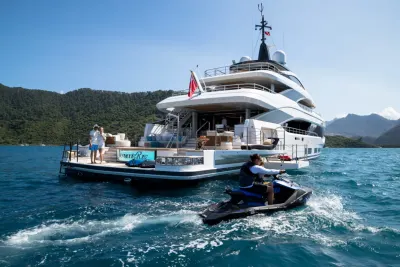 A person rides a jet ski in the foreground with a large yacht anchored in the background, surrounded by lush green hills under a clear blue sky.