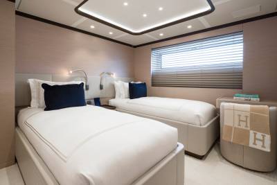 Two neatly made twin beds with navy pillows occupy the foreground, while a window with blinds and a small side table with a blanket and books form the background.