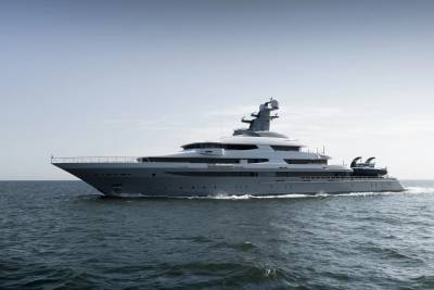 A sleek, gray superyacht cruises on calm, dark blue water under a pale, hazy sky.