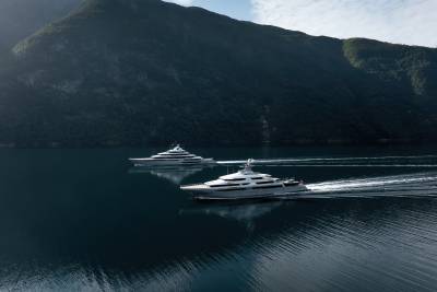 Two sleek yachts glide across a calm, dark blue body of water, leaving white wakes, with a majestic, tree-covered mountain range rising in the background under a hazy sky.