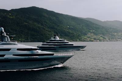 Two large yachts sail on calm water in the foreground, with a lush green mountainside dotted with houses in the background.