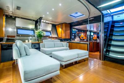 A modern, light-colored sectional sofa and ottomans fill the foreground of a luxurious yacht interior, while the background reveals a sleek galley and a curved staircase.